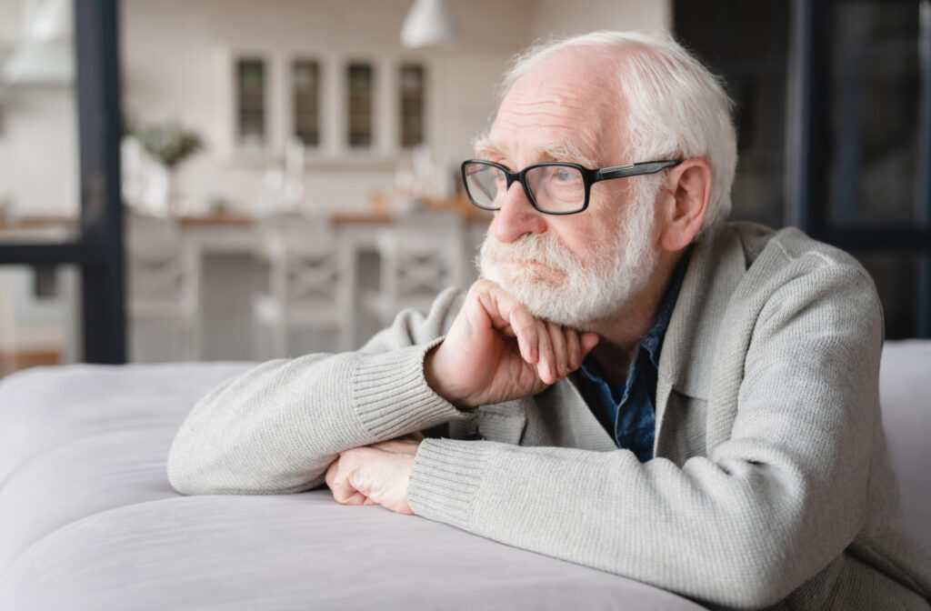 An older man with glasses sitting on a couch while gazing out of a window