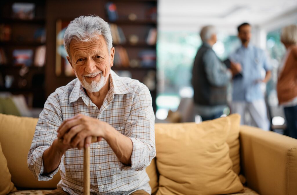 A senior man sitting on the couch at an assisted living community with other residents chatting in the background