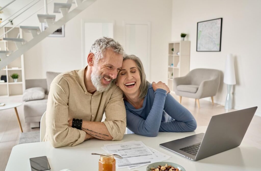 An older adult leans on their spouses shoulder and laughs while they research communities together in their kitchen
