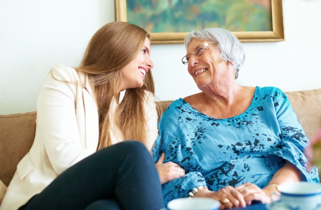 An adult child sits and clasps their older parent's arm while they share a laugh on a couch in assisted living
