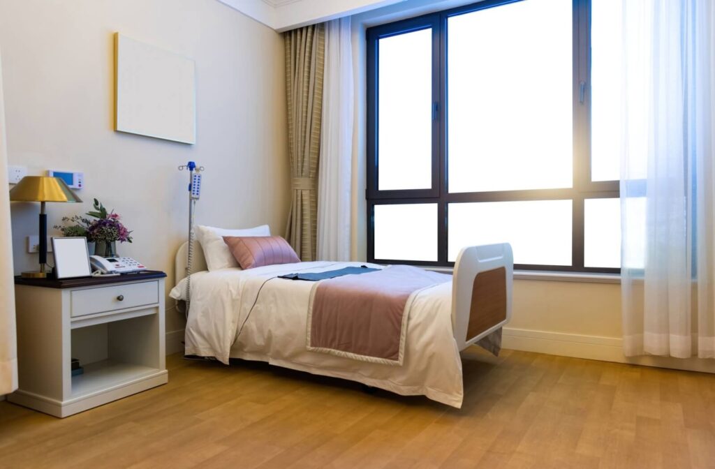 A clean bedroom in skilled nursing, with a well-made bed and nightstand resting underneath a brightly lit window